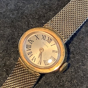 May include: A gold-toned Timex wristwatch with a silver face and Roman numeral hour markers. The watch has a mesh metal band.