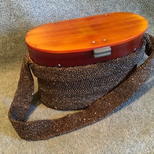 Puede incluir: Bolso vintage ovalado con cuerpo y correa de cuentas. La parte superior es de color marrón rojizo con una tapa ovalada marrón más clara. El bolso tiene un cierre plateado. Un accesorio clásico de mediados del siglo XX.