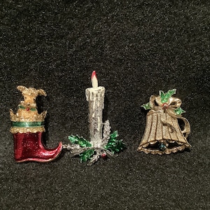 May include: Three vintage Christmas brooches featuring a stocking, a candle, and a bell. The stocking is red with a green cuff and a gold crown. The candle is white with a red flame and green holly leaves. The bell is gold with a green bow and holly leaves.