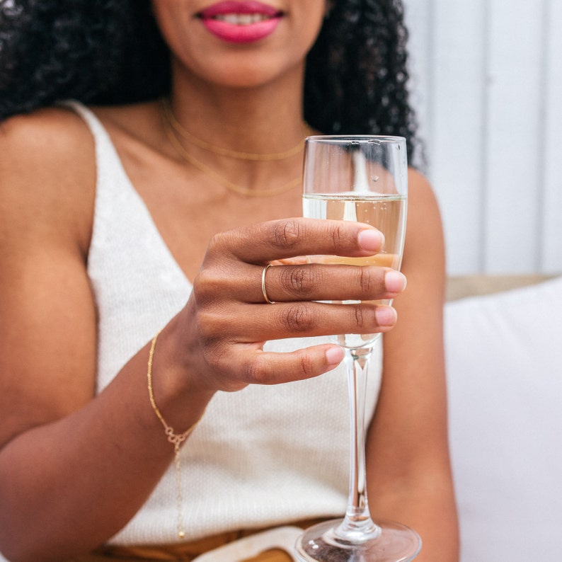 Może przedstawiać: Kobieta w białej koszulce bez rękaw&oacute;w trzyma kieliszek szampana. Ma na palcu złoty pierścionek.