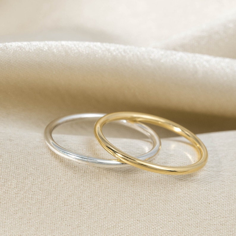 Może przedstawiać: Dwa proste obrączki ślubne, jedna złota, a druga srebrna, leżą na sobie na kremowym tle z tkaniny.