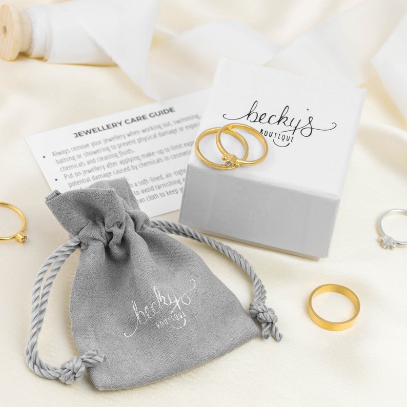 Może przedstawiać: Szary aksamitny woreczek na biżuterię z napisem "beckyo" i słowami "with love" mniejszą czcionką. Woreczek leży na białym tle obok białego pudełka z napisem "becky's BOUTIQUE" na g&oacute;rze. W pudełku znajdują się dwa złote pierścionki.