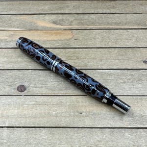 May include: A stylish pen with a blue and brown leopard print pattern. The pen has silver accents and a silver tip. The pen is lying on a wooden surface.