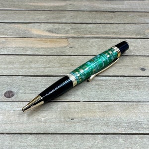 May include: A ballpoint pen with a unique design. The pen features a green circuit board pattern on the barrel, complemented by gold-colored accents and a black cap. The pen has a gold-colored tip and a gold-colored clip.