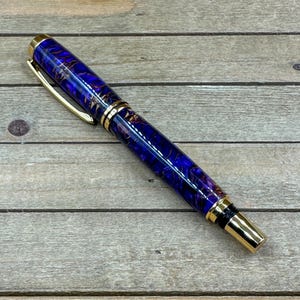 Puede incluir: Una pluma estilográfica artesanal con un vibrante barril de resina azul y dorado. La pluma presenta detalles dorados en el clip, el plumín y los adornos. La pluma se exhibe sobre una superficie de madera.