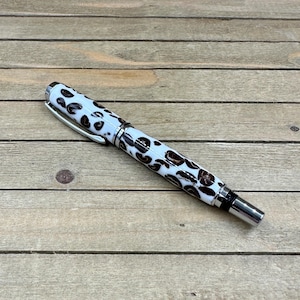 May include: A white pen with a brown spotted pattern and silver accents. The pen has a silver clip and a silver tip. The pen is lying on a wooden surface.