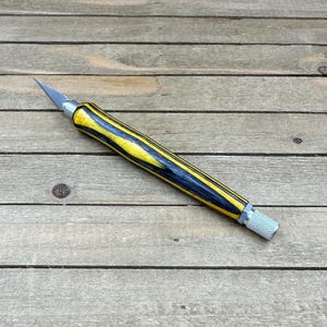 May include: A craft knife with a yellow and black striped wooden handle and a silver blade. The handle has a textured grip at the end. The knife is designed for precision cutting tasks.
