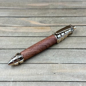 Puede incluir: Un bolígrafo artesanal con un cuerpo de madera oscura y detalles metálicos plateados. El bolígrafo tiene una punta y un clip. El bolígrafo se muestra sobre una superficie de madera.