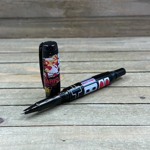 May include: Black pen with a cap featuring a pixelated image of Mario from Super Mario Bros. The pen's barrel has the words "SELECT START" and a red and white design. The pen is on a wooden surface.