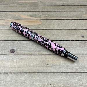 May include: A pink and black leopard print pen with silver accents. The pen has a sleek, modern design and is made of a glossy material. The pen is a writing instrument and is suitable for everyday use.