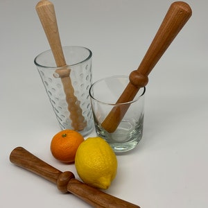 May include: Wooden muddlers in a variety of shades, with a light-colored wood and a darker, reddish-brown wood. Two clear glasses and a lemon and orange are also in the image. The muddlers are designed for mixing drinks.