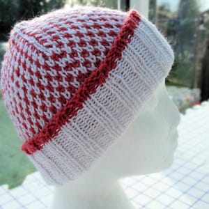 Puede incluir: Un gorro de punto blanco y rosa con ribete rojo. El gorro está hecho de un hilo suave y texturizado.