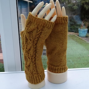 Puede incluir: Guantes sin dedos de punto amarillo mostaza. Uno presenta un diseño de punto trenzado, el otro un patrón de punto más sencillo. Los guantes se exhiben en manos de maniquí, mostrando la textura y el color de los accesorios.