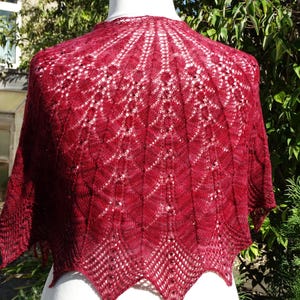 May include: A burgundy-colored shawl with a delicate, openwork design. The shawl is draped over a mannequin, showcasing its intricate knitted pattern. The edges have a scalloped trim, adding a touch of elegance to this handmade accessory.