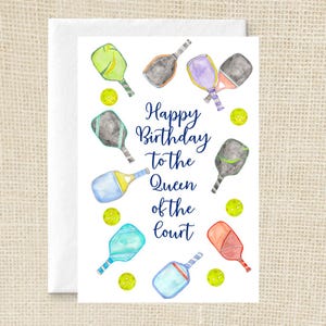 May include: A white greeting card featuring watercolour illustrations of various colourful pickleball paddles and yellow pickleballs surrounding the dark blue text that reads Happy Birthday to the Queen of the court in a script and print font combination.
