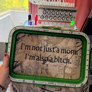 以下が含まれることがあります： 「I'm not just a mom, I'm also a bitch.」のテキストが書かれた迷彩柄の生地を保持する緑色の長方形の刺繍枠。背景には刺繍ミシンがあります。
