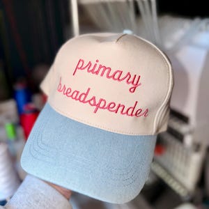 Könnte beinhalten: Beige und hellblaue Baseballkappe mit dem gestickten Text "primary breadspender" in Rosa. Die Kappe hat einen gebogenen Schirm und eine strukturierte Krone. Der Hintergrund zeigt eine Stickmaschine.