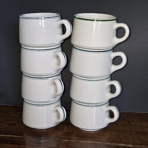 May include: Eight white ceramic mugs stacked in two columns of four. Each mug features a simple, thin dark green horizontal band encircling the upper portion of the body and a matching green stripe along the outer edge of the curved handle.
