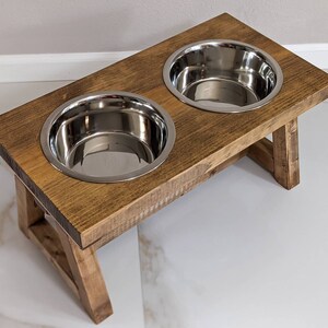 May include: A wooden elevated dog bowl stand with two stainless steel bowls. The stand is stained a dark brown color and has a rustic look.