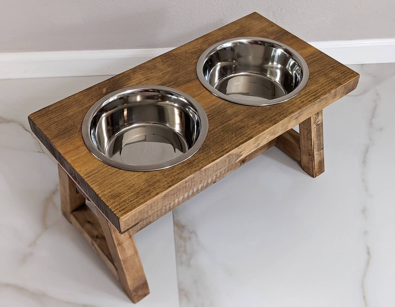 May include: A wooden elevated dog bowl stand with two stainless steel bowls. The stand is stained a dark brown color and has a rustic look.