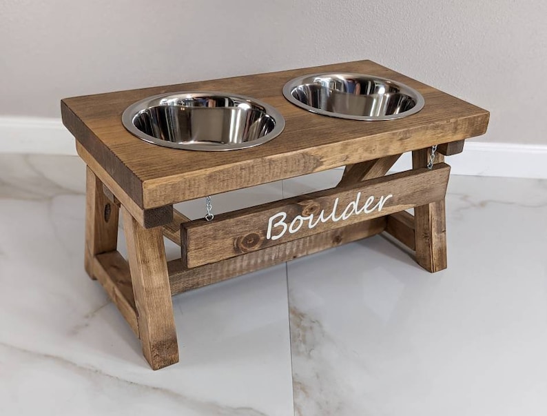 May include: A wooden dog bowl stand with two stainless steel bowls. The stand is stained brown and has the name "Boulder" engraved on the front.