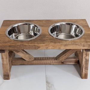 May include: A rustic wooden dog bowl stand with two stainless steel bowls. The stand has a distressed brown finish and a simple, farmhouse style.