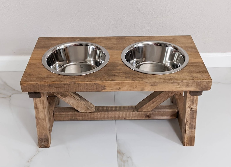 May include: A rustic wooden dog bowl stand with two stainless steel bowls. The stand has a distressed brown finish and a simple, farmhouse style.