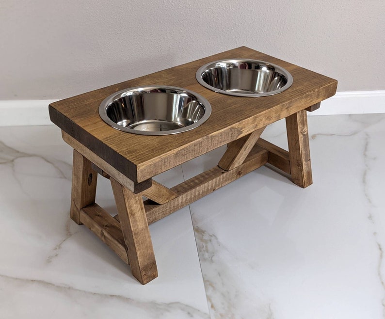 May include: A rustic wooden dog bowl stand with two stainless steel bowls. The stand has a distressed brown finish and a farmhouse style.
