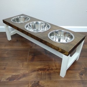May include: A wooden pet food stand with three stainless steel bowls. The stand is painted white and has a dark brown wood top.