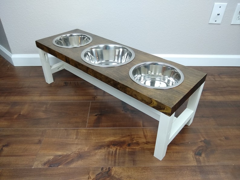 May include: A wooden pet food stand with three stainless steel bowls. The stand is painted white and has a dark brown wood top.