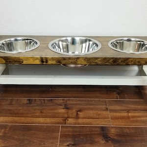 May include: A wooden dog food stand with three stainless steel bowls. The stand is painted white and has a brown wooden top.