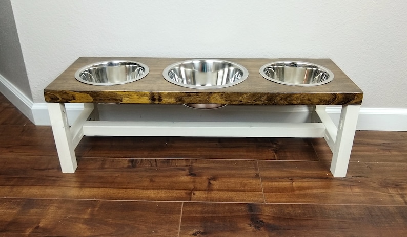 May include: A wooden dog food stand with three stainless steel bowls. The stand is painted white and has a brown wooden top.