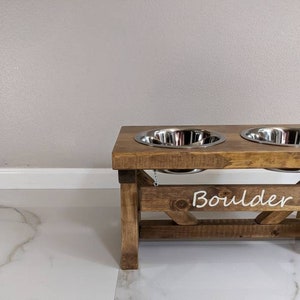 May include: A wooden dog bowl stand with two stainless steel bowls. The stand is stained brown and has the word "Boulder" written on the front.