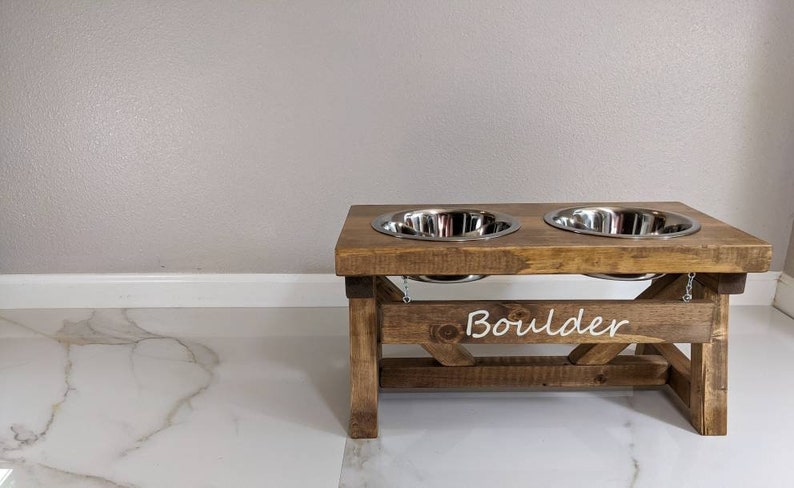 May include: A wooden dog bowl stand with two stainless steel bowls. The stand is stained brown and has the word "Boulder" written on the front.