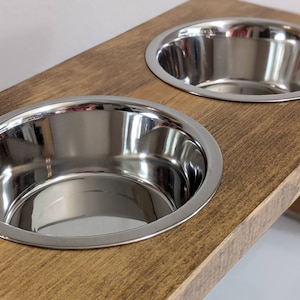 May include: A wooden elevated dog bowl stand with two stainless steel bowls. The stand is stained a dark brown color.