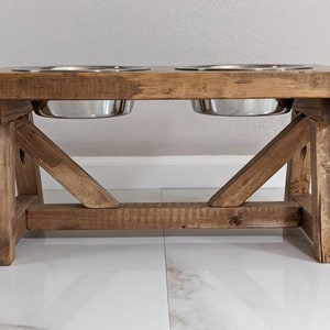 May include: A rustic wooden dog bowl stand with two stainless steel bowls. The stand has a brown stain and a simple, farmhouse style.