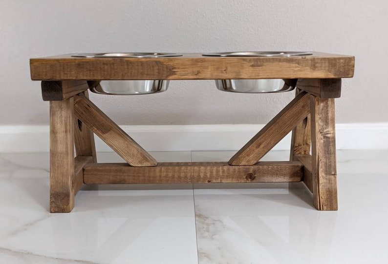 May include: A rustic wooden dog bowl stand with two stainless steel bowls. The stand has a brown stain and a simple, farmhouse style.