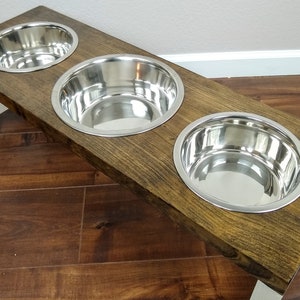 May include: A wooden dog food stand with three stainless steel bowls. The stand is stained a dark brown color and has white legs.