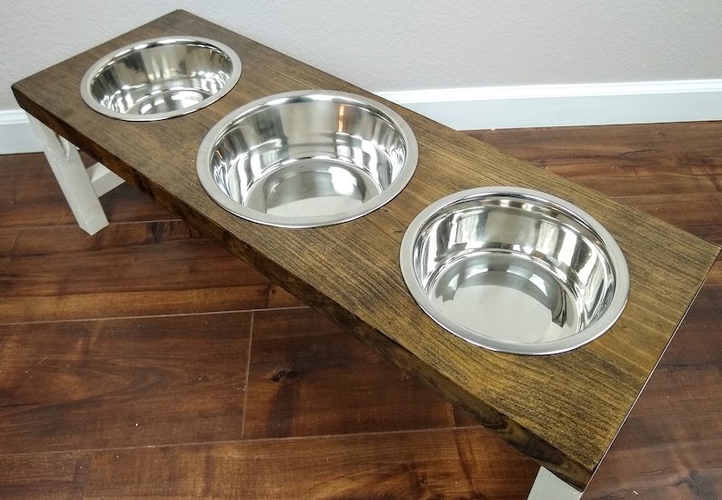 May include: A wooden dog food stand with three stainless steel bowls. The stand is stained a dark brown color and has white legs.