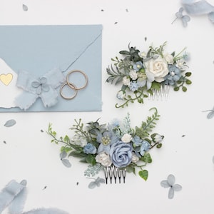 Op de afbeelding: Twee blauwe en witte bloemenhaar kammen met zilveren kammen. De kammen liggen op een witte achtergrond met een blauw lint en gouden ringen.