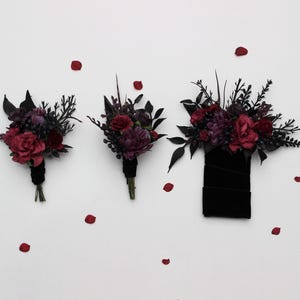 以下が含まれることがあります： 白背景に、濃い赤と紫の花と黒のアクセントが施された3つのフラワーアレンジメントが表示されています。アレンジメントには、ブートニアと大きな花束が含まれており、赤い花びらが散らばっています。花束は黒いリボンで結ばれています。