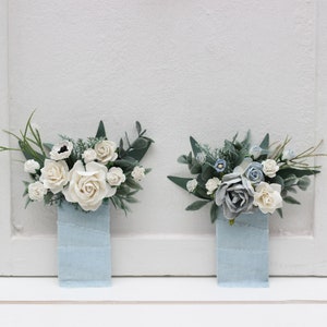 May include: Two small bouquets of artificial flowers in blue and white colors, arranged in blue fabric pouches. The bouquets are made of white roses, blue hydrangeas, and green leaves.