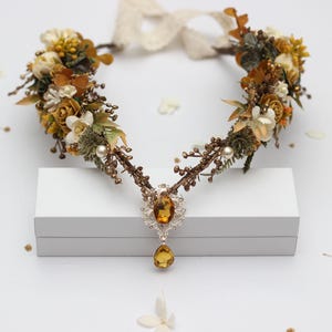 Puede incluir: Una corona floral con diseño en forma de V, con una mezcla de flores amarillas, blancas y marrones, acentuada con perlas y un gran colgante de joya color ámbar. La corona se encuentra sobre un fondo blanco.