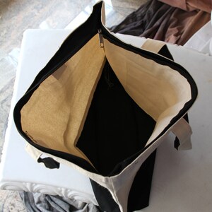 May include: A black and white canvas tote bag with a zipper closure. The bag is open and the inside is visible. The bag is made of a durable canvas material.