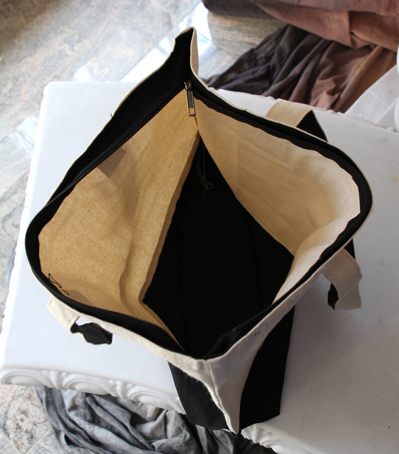 May include: A black and white canvas tote bag with a zipper closure. The bag is open and the inside is visible. The bag is made of a durable canvas material.