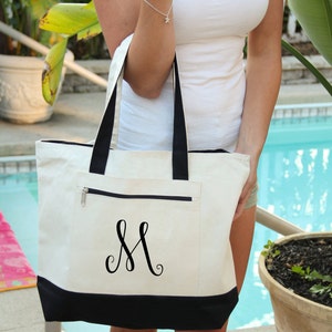May include: A white canvas tote bag with black trim and a black embroidered letter "M" on the front. The bag has a zippered pocket on the front.