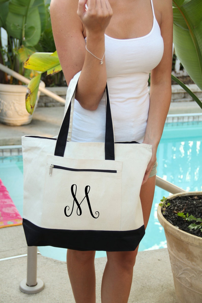 May include: A white canvas tote bag with black trim and a black embroidered letter "M" on the front. The bag has a zippered pocket on the front.