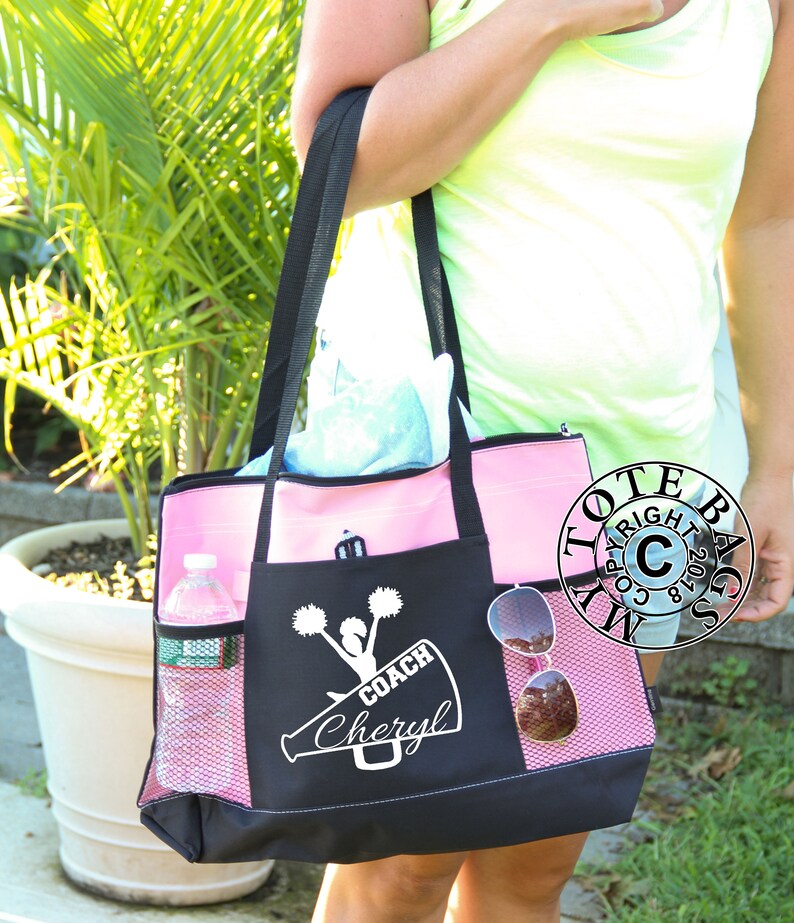 May include: Black and pink tote bag with a white design of a cheerleader holding pom-poms and a megaphone. The text "COACH Cheryl" is on the megaphone.