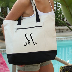 May include: A white canvas tote bag with a black bottom and a black zippered pocket. The bag has a black monogram of the letter "M" on the front.
