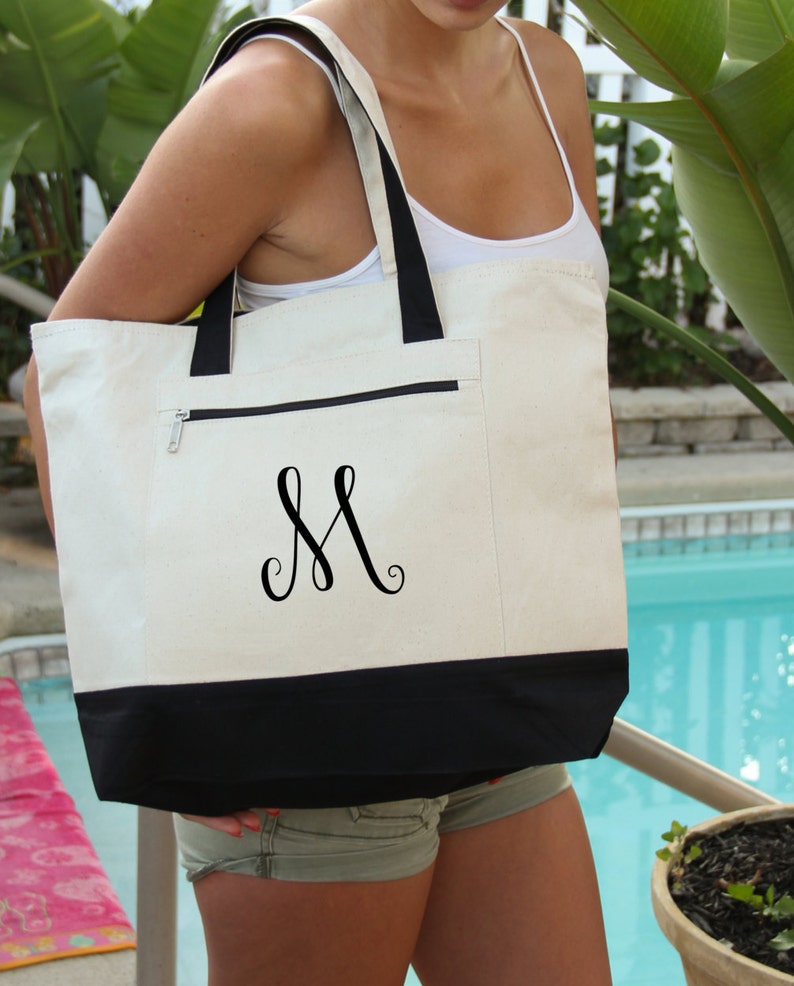 May include: A white canvas tote bag with a black bottom and a black zippered pocket. The bag has a black monogram of the letter "M" on the front.